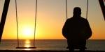 A man is sitting alone on a swing looking at the sunset.