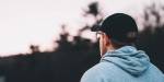 A lonely man wearing a hoodie and a baseball cap, seen from behind, as he gazes towards the evening sky.