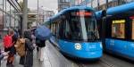 Photo of trams at the stop Holbergs plass in Oslo. Passengers with umbrellas stand ready to board the tram.