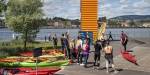 Gruppe mennesker i redningsvester står på land foran Oslofjorden og ser på en instruktør med kayak. Bak dem ligger det mange fargerike kayaker.