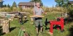 Kyrre Andersen out in the yard, surrounded by logs, wood materials and various machines. He shows off a piece of wood. In the background we see a small red house.