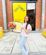 Carla Louise Hughes smiling with a diploma and a bouquet of flowers in front of a OsloMet building.