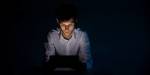 A young man sits in front of a computer in a dark room, only the screen lighting him up.