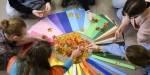 Several people sit in a ring on the floor, bending over some colored sheets and leaves in autumn colors, located in the center of the ring.