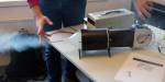 The smoke machine blows smoke out of the wind tunnel, which is mounted on a table. One of the students starts the machine and sticks one hand into the smoke that comes out of the machine.