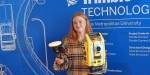 Picture of newly graduated bachelor's student in civil engineering, Cecilie Erland, with equipment for the lab in her hands. The background is blue with drawings of roads, vehicles, lampposts and other transport infrastructure, and information about Trimble's solutions.