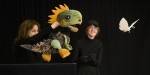 To drama- og teaterstudenter i svarte klær fremfører dukketeater på en scene. Den ene studenten holder opp en dinosaurdukke, mens den andre holder to svarte stenger hvor en papirsommerfulg er festet i enden.