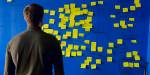 Man stands with his face towards a big blue wall with many yellow post-it notes.