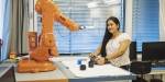 Smilende kvinnelig student jobber med en stor oransje industrirobot, i noe som ligner et klasserom.