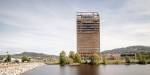 Mjøstårnet som er bygd i kryslaminert tre. Vann fra Mjøsa i forgrunnen, lettskyet himmel og lave åser i bakgrunnen. Parkområde med benker til venstre.
