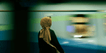 a young woman in a head scarf with her back to the camera and a train moving behind her