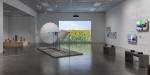 An exhibition from Nitja Centre for Contemporary art. A big room with white walls and concrete floor. There are monitors and pictures on one wall, a projector shows a picture of sunflowers on another. In the middle of the room stands an artwork with stands and umbrellas.
