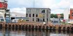 The image shows the facade of Oceanlab at Filipstadkaia quay. The building has impregnated timber walls and windows that stretch almost from floor to ceiling. OsloMet’s logo is on the wall. In the foreground is the quay with car tyres used as dock fenders, and the ocean. Winches and other buildings on the quay can also be seen in the image. The sky is partly cloudy.