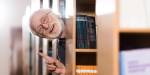 Eldre mann med skjegg stikker hodet og pekefinger ut mellom bokreolene på bibliotek