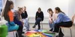 Six people sit in a semicircle on separate chairs. Everyone looks down the middle where there are colored sheets and autumn leaves.