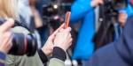 A smartphone is held in the middle of a media crowd, seemingly filming an event.