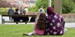 Mor og datter sitter sammen på gresset i en park med ryggen til. I bakgrunnen sitter og står flere andre personer.