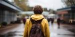 Ung gutt med gul jakke og brun ryggsekk med ryggen til foran en skole