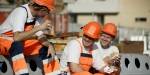 Tre arbeidere i oransje arbeidsklær og hjelm spiser matpakka si ute i sola på en byggeplass.