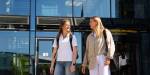 Two students in sunny weather in front of a modern building.