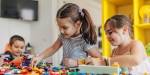 To jenter og en gutt smiler og leker med fargerike legoklosser ved et bord i klasserommet.