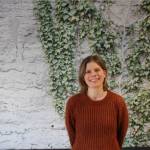 Portrettbilde av forsker Hilde Zeiner som står foran en grå vegg med grønn eføy og ser i kamera og smiler.