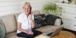 Elderly woman sitting on a couch with an open laptop and a paper form in front of her while talking on the phone.
