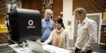 Picture of Maryam showing something on the quantum computer to professors Sergiy and Sølve who are standing on opposite sides of her. On the table in front of them is a black quantum computer