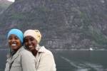 To smilende jenter med en fjord, en ferge og bratte fjellsider i bakgrunnen.