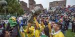Fotballspillere med gule drakter foran Rådhuset løfter en pokal