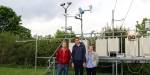 Bilde av de tre studentene  i hagen til Meteorologisk institutt. I bakgrunnen er det et stillas med måleutstyr.  Det er sommer og grønt på trærne, og det er overskyet himmel i bakgrunnen.