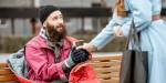 En mann med skjegg og svart lue sitter på en parkbenk med en rød sovepose rundt seg og får en kaffekopp av en dame i blå kåpe med svart veske over armen.