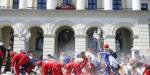 Norwegian royal palace facade with the royal family on the balcony and graduating high schoolers walking and crawling on the ground in a parade dressed in red or blue "russ" outfits.