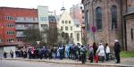 People standing in line on a sidewalk in a Norwegian city. Many carry empty shopping bags.