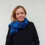 NIBR-forsker Heidi Bergsli i svart kåpe og blått skjerf mot en hvit vegg ser i kamera og smiler.