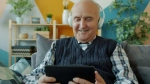 An elderly man is sitting on a sofa wearing headphones and looking at a tablet.