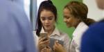 To mørhårede 16-årige jenter ser sammen på mobiltelefon og smiler.