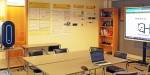 Picture from the quantum lab, with one of the quantum computers on the left, posters presenting the research project on the wall, and a bookshelf and screen on the right. In the foreground a table with a laptop.