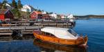 Uthamn i Merdø i Agder, kystripe med kvite og raude hus langs sjøen med ein brun trebåt som ligg fortøydd til ei bryggje.