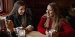 Two female students in thick sweaters holding around their cups of hot chocolate at a cafe.