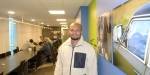 Photo of Faisal in the hallway outside the classrooms. There are workplaces for students on the left, and a group of students are sitting in the background. On the right there are pictures on the wall.