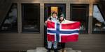 To personer står foran et trehus og holder et norsk flagg. Huset har tre synlige vinduer, og det ligger snø på bakken til venstre i bildet.