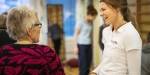 Helsevitenskap-student i hvit uniform snakker og ler med en eldre kvinne.