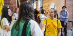 En jente med gul OsloMet t-skjorte hilser på en annen student som står med ryggen mot kamera. Jenta smiler.