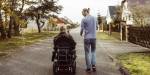 A man in a wheelchair and a standing man walk down a suburban street, bordered by trees and houses.