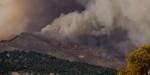 Image of dark and heavy clouds lying low over a mountain. Sun over forest and settlement below, but the clouds warn of a change in weather.