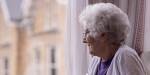 Eldre kvinne sitter alene inne og ser ut av et vindu.
