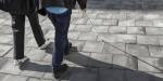 Viser beina og den hvite stokken til en blind person som går på et grått, hellelagt fortau. Ved siden av han går det en person i følge med han.