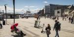 A big open plaza, modern buildings on one side and a ferry on the water on the other side. People strolling and neatly parked el scooters.