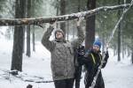 To menn med luer på hodet i en skog dekket med noe snø, begge løfter en stor stokk hver